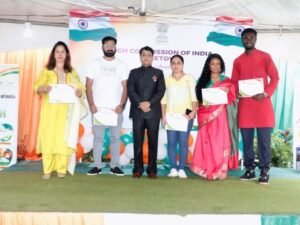 Six young adults on a stage holding certificates, with Indian flags and a government banner behind them.
