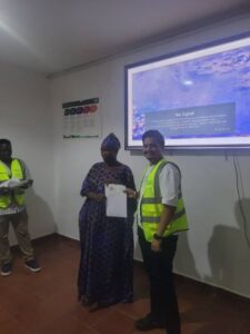Man in a safety vest hands a certificate to a woman in a blue dress inside a room with a projector screen; another person stands nearby.