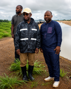 Three men stand on a dirt road beside a narrow, water-filled canal; one wears a reflective black jacket and beige cap, another in a dark blue outfit, with a third man behind them as vehicles appear in the background.