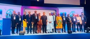 Group of diverse professionals on a conference stage posing for a photo with lanyards and badges visible on everyone present, blue backdrop behind them.