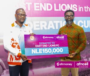 Two men hold a large purple donation check reading 'DONATED TO EAST END LIONS FC' with logos 'afrMoney' and 'Africell'; banner behind them shows bold white text and pink-to-blue gradient.