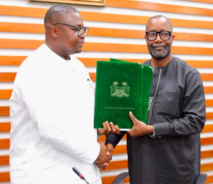 Two men shake hands while one hands over a green folder with the Sierra Leone crest, in a formal setting.
