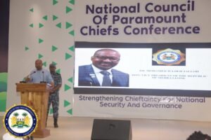 Speaker at a wooden podium addressing the National Council of Paramount Chiefs Conference, with a uniformed guard beside him on stage.
