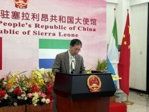 | The Calabash Newspaper Man in a suit giving a speech at a podium with the Chinese emblem, in front of a banner and flags.