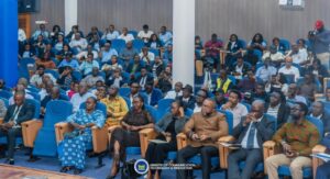 | The Calabash Newspaper Large audience seated in blue chairs at a conference hall, many people watching a presentation.