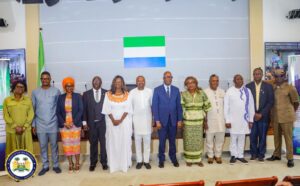Group of dignitaries and officials standing in a row at a formal event, with a green-white-blue flag behind them.