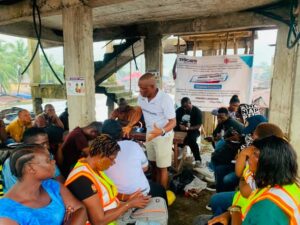 | The Calabash Newspaper A man in a white shirt and shorts stands and speaks to a group seated under a concrete shelter; several attendees wear orange safety vests, listening attentively as a banner with logos hangs in the background.