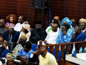 | The Calabash Newspaper Audience of diverse people seated in a formal event, some wearing traditional African attire and suits.