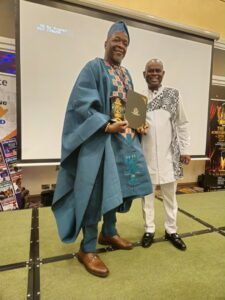 | The Calabash Newspaper Man in blue traditional outfit receiving an award on stage, holding a plaque and smiling, beside a casually dressed man in white and black patterns on an event backdrop.