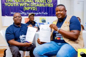Two men sit on white chairs at a ceremony, each holding a document and smiling; a purple banner behind them reads 'The National Panel of Youth with Disabilities (NPYD)'.