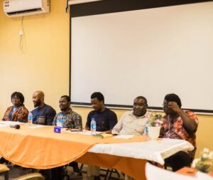 | The Calabash Newspaper Six men sit as a panel at a long table with an orange tablecloth, water bottles and a blank projector screen behind them in a meeting room.