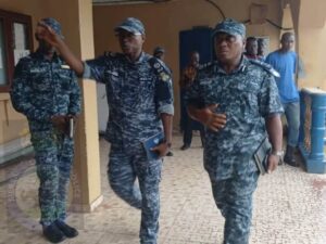 Three people in blue digital camouflage military uniforms walk through a tiled hallway; two hold tablets/notebooks, and the front-right person gestures as they move. A fourth person in civilian clothing looks on in the background.