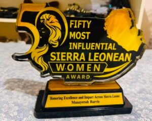 | The Calabash Newspaper Gold and black trophy in the shape of Sierra Leone with bold text reading 'Fifty Most Influential Sierra Leonean Women Award' and a plaque honoring Musayoreh Barrie on a table.