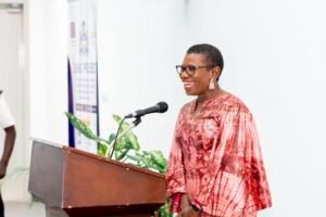 Smiling woman in a pink patterned dress stands at a wooden podium with a microphone, delivering a speech.