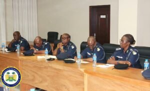 Five police officers in uniform sit at a long wooden conference table during a formal meeting, with water bottles and caps on the table, and a government seal in the corner.