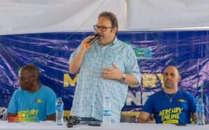 Man center speaks into orange microphone at a panel, flanked by two men in blue shirts with water bottles on the table. Banner behind reads Mercury Online.