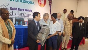 Man in a suit hands a wrapped package to a student in a white uniform at a scholarship ceremony; banner reads 'Baba Guru Nanak Scholarships' in the background.
