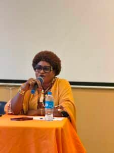 African woman in an orange outfit speaks into a microphone at a panel table, with a water bottle and papers in front of her.
