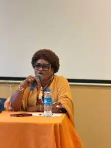 African woman in an orange outfit speaks into a microphone at a panel table, with a water bottle and papers in front of her.