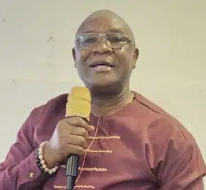 Monk in maroon robes holding a yellow microphone, wearing glasses and a beaded bracelet.