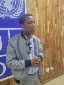 UNDP Lesotho Deputy Resident Representative, Dr. Taye Amssalu | The Calabash Newspaper Man in a gray jacket speaks in front of a blue United Nations banner, against a wooden wall backdrop.