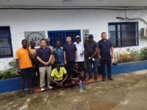Group of men posing for a photo outside a white building with blue window bars; signs reference a fisheries organization or office.