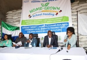 Five panelists seated at a long table during a Home-Grown School Feeding event, with a large banner about the program in the background.
