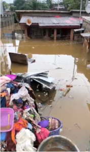 Residential street flooded with muddy water covering ground floor, debris and belongings piled along the curb near a damaged house.