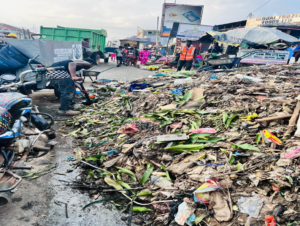 Trash and debris piled along a market street, with a worker in an orange vest nearby inspecting the pile.