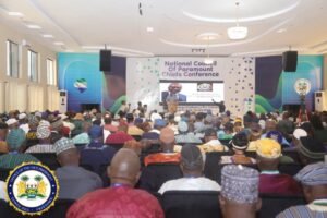 Large conference hall filled with attendees watching a speaker at a podium during the National Council Of Paramount Chiefs Conference; stage lights and screens behind.