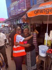 Two women hug and smile in a bustling street market, one wearing an orange safety vest among colorful stalls and umbrellas.