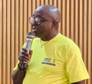 Man with glasses speaks into a microphone, wearing a bright yellow Amnesty International shirt, with a wooden slat backdrop.