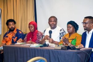 | The Calabash Newspaper Panel of five speakers at a conference, with one person speaking into a microphone and others seated with notebooks at a long table on stage.