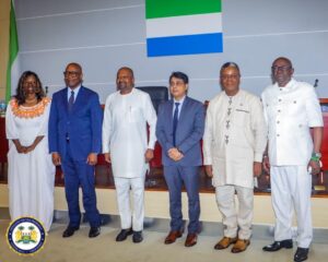 Six officials in formal attire pose for a group photo on a stage with the Sierra Leone flag behind them, government setting.