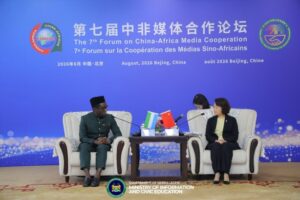 Two officials seated on a stage in cream armchairs, facing each other, with small table between them holding flags (China and Sierra Leone) during The 7th Forum on China-Africa Media Cooperation; a blue backdrop displays the event title in Chinese and English and government logos at the bottom.