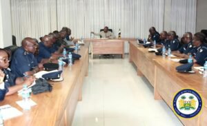 | The Calabash Newspaper Formal meeting of uniformed officers seated along U-shaped wooden conference tables, with water bottles and notes; government seal visible in the lower right corner