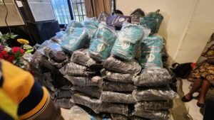Large stack of bundled bags wrapped in plastic on the floor in a room, with people and windows in the background.