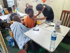 Health worker in a black shirt examines a man in an orange shirt at a crowded clinic, others seated at tables nearby.