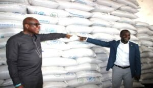 Two men in a warehouse fist-bump in front of tall stacks of white bags, signaling a partnership or deal.