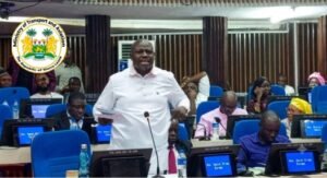 | The Calabash Newspaper Man in a white shirt speaks from a podium in a conference room filled with seated attendees; Ministry of Transport and Communications logo visible in the top left.