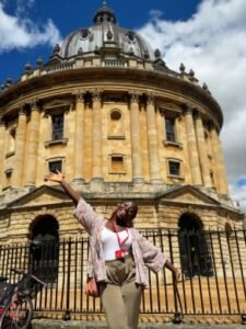 | The Calabash Newspaper Woman wearing a patterned blouse and beige pants takes a selfie in front of a large round-domed stone building with columns, behind a black fence, with a bicycle nearby.