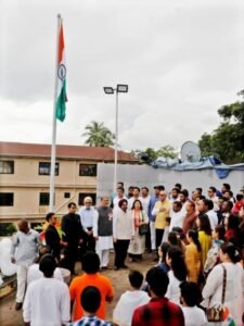 Diverse group of people on a rooftop terrace watching a flag-raising ceremony as the Indian flag waves on a tall pole, with buildings in the background.