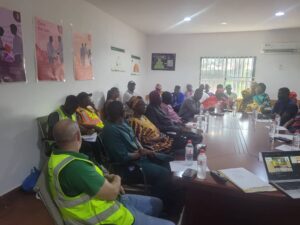 A large meeting room with about 25 people seated around a long conference table, some wearing safety vests, listening to a presentation.