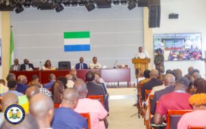 | The Calabash Newspaper Panel of officials seated at a long table on a stage, speaker at a podium addressing the audience with the Sierra Leone flag behind them.
