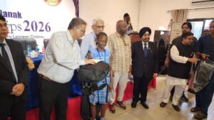Young girl in blue dress receives a backpack from a man in a striped shirt at a formal ceremony; others stand nearby near a banner reading 2026.