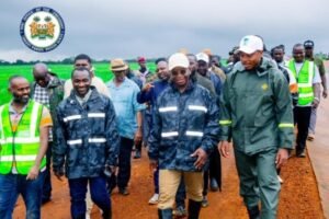 Group of officials and workers walking along a dirt road at a road construction site; the Office of the Prime Minister seal is in the top-left corner.