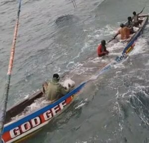 | The Calabash Newspaper A long, narrow rowing boat with several men paddling in rough, choppy water; side of the hull is blue with red and white lettering that reads 'GOD GEF'
