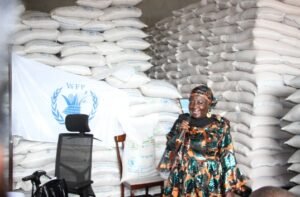 | The Calabash Newspaper Woman in a colorful dress speaks into a microphone at a World Food Programme event, with stacked sacks behind her and a WFP banner visible on the left.
