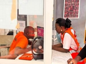 | The Calabash Newspaper Inmate in an orange jumpsuit talks with a staff member in a high-visibility vest at a glass service window; a desk fan sits on the counter.