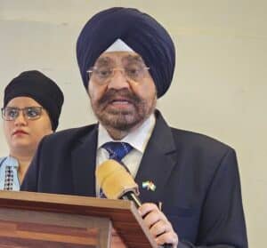 Man in a dark suit, blue turban, and glasses speaks at a wooden podium with a microphone; small flag pin on lapel is visible.
